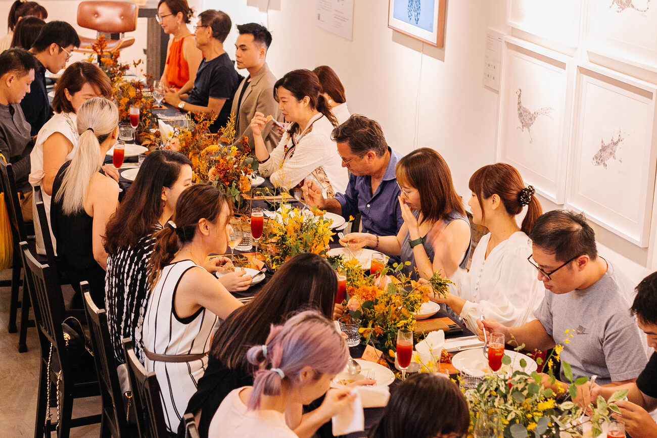 A long table of guests eating at the Autumn Feast dinner, framed drawings on the white wall behind them