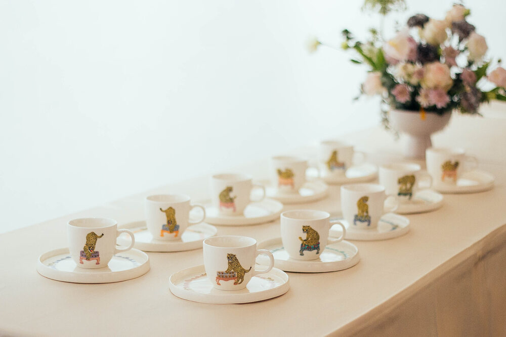 A row of illustrated ceramic cups and saucers arranged on a white table with flowers