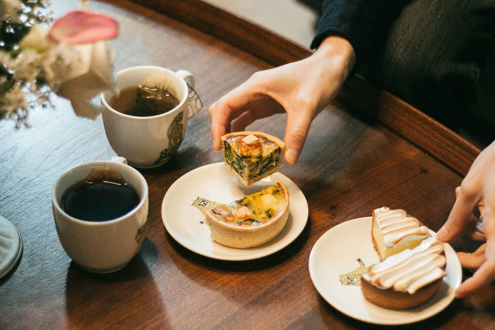 Hands reaching for tarts and tea in illustrated cups on a wooden table