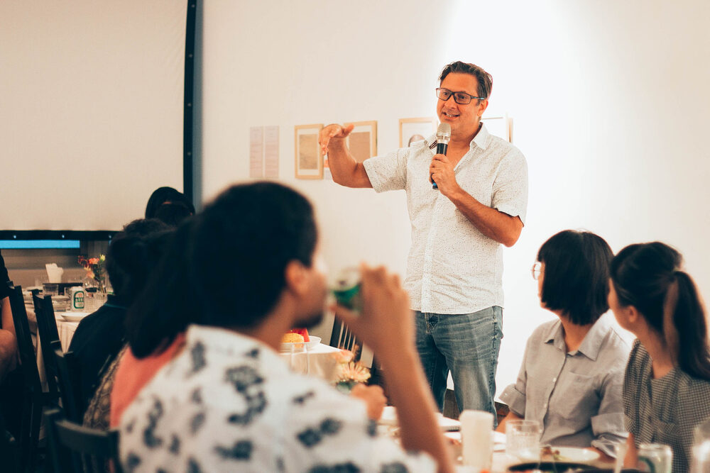 The artist speaking into a microphone to seated guests in front of a hung wall of works